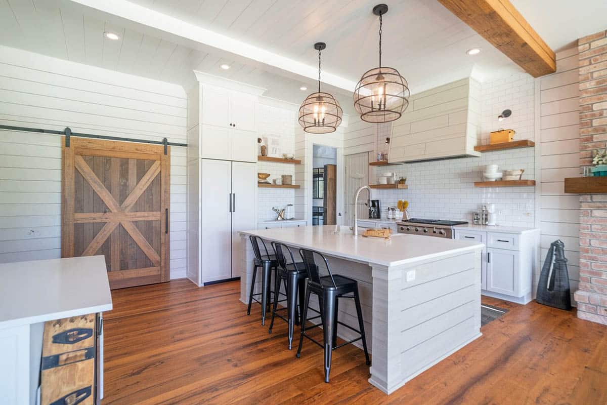 Beautiful kitchen with cabinetry in bright white, shiplap surround island, wood beam ceiling, sliding barn doors and open wooden shelving